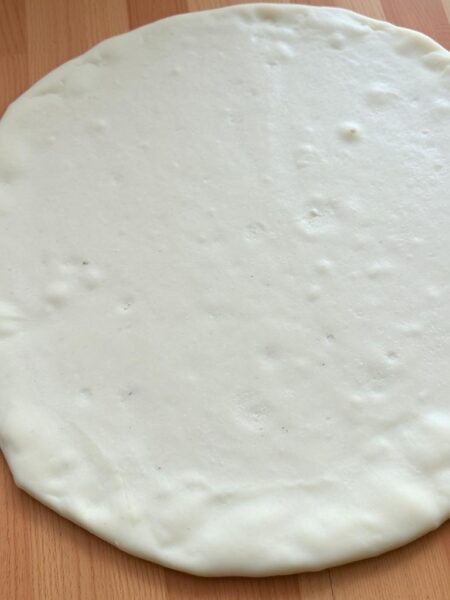 A flat, thinly rolled-out pizza dough on a wooden surface. The dough has a light, airy texture with a few small air bubbles visible. The surface is slightly mottled, with a soft, off-white color. The lighting is soft and natural, creating subtle shadows that accentuate the dough's delicate structure. The camera angle is slightly elevated, providing a clean, uncluttered view of the dough. The overall mood is one of simplicity and homemade comfort, inviting the viewer to imagine the possibilities of this versatile canvas for flavorful toppings.