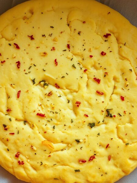 A freshly baked, golden-brown pizza dough, its surface dotted with flecks of minced garlic and fragrant herbs. The dough's texture is slightly chewy, with a faint aroma of yeast wafting through the air. The camera angle captures the dough from an overhead perspective, highlighting its circular shape and the intricate patterns created by the ingredients. Soft, natural lighting casts a warm glow, accentuating the dough's inviting appearance. The background is slightly blurred, allowing the pizza dough to take center stage and entice the viewer to imagine the delicious possibilities of this flavorful foundation for a homemade pizza.