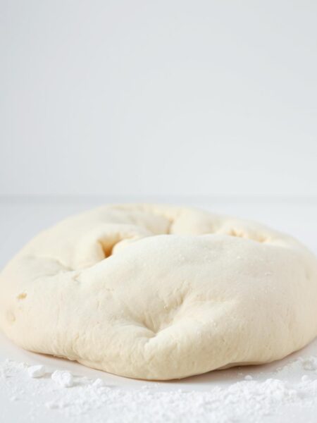 A pristine white surface, bathed in soft, diffused lighting, showcases a classic all-purpose flour pizza dough, its smooth, supple texture glistening invitingly. The dough is artfully arranged, its edges slightly curled, revealing a subtle yeasty aroma. In the foreground, a few scattered flour granules add a rustic touch, while the background maintains a clean, minimalist aesthetic, allowing the dough to take center stage. The image conveys a sense of simplicity, highlighting the humble yet essential nature of this foundational ingredient for homemade pizza.
