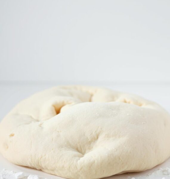 A pristine white surface, bathed in soft, diffused lighting, showcases a classic all-purpose flour pizza dough, its smooth, supple texture glistening invitingly. The dough is artfully arranged, its edges slightly curled, revealing a subtle yeasty aroma. In the foreground, a few scattered flour granules add a rustic touch, while the background maintains a clean, minimalist aesthetic, allowing the dough to take center stage. The image conveys a sense of simplicity, highlighting the humble yet essential nature of this foundational ingredient for homemade pizza.