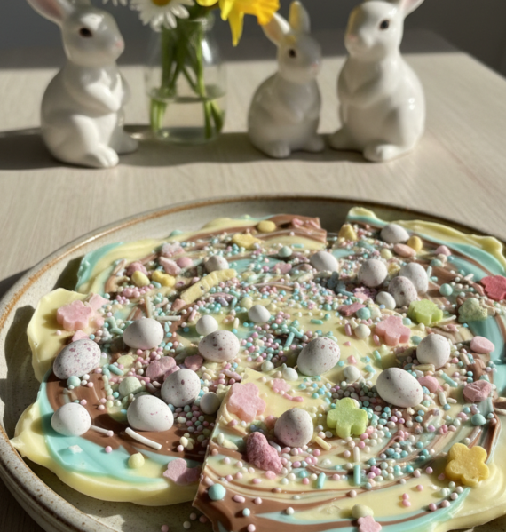 A beautifully arranged plate of vibrant Easter Candy Bark sits prominently in the foreground, showcasing a colorful variety of melted chocolate with swirls of pastel colors. The bark is generously sprinkled with an assortment of mini eggs, sprinkles, and crushed candies, adding texture and visual interest. In the middle, a soft-focus background features a light wooden table surface adorned with spring-themed decorations such as small flowers and bunnies, enhancing the festive atmosphere. Natural light streams in from the left, casting gentle shadows that highlight the bark's glossy surface. The overall mood is cheerful and inviting, perfect for celebrating Easter with family and friends. The image captures the essence of joy and creativity in holiday baking.